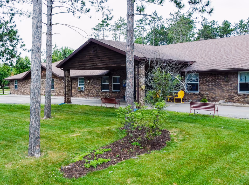 Brick single-story senior living building with a covered entrance, benches, trees, and a grassy lawn.