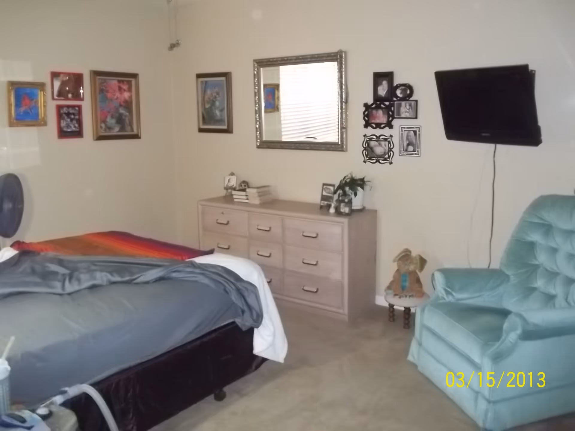 A bedroom with a bed covered in gray and multicolored blankets, a light wooden dresser with books and plants on top, a large mirror above the dresser, several framed pictures on the walls, a mounted flat-screen TV, and a light blue upholstered armchair next to a small round table with a stuffed bunny on it.
