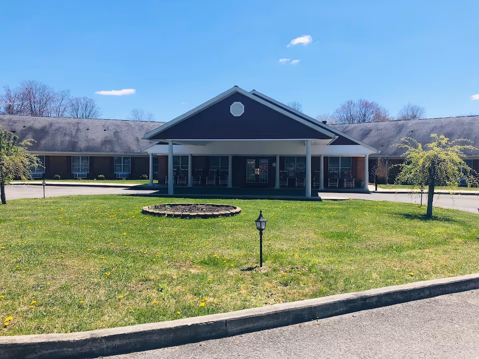 Front exterior view of Mountain Memories Assisted Living & Retirement Center building with a covered entrance, rocking chairs on the porch, a circular flower bed, small trees, and a grassy lawn under a clear blue sky.