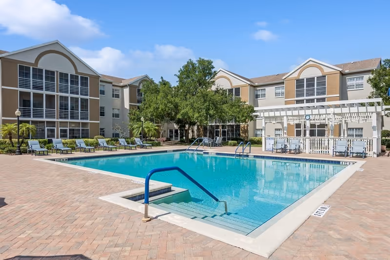 Outdoor swimming pool with clear blue water surrounded by a brick-paved deck. Several lounge chairs are lined up along the left side of the pool. Behind the pool are two three-story buildings with large windows and screened balconies. There is a white pergola structure on the right side near the pool with some chairs underneath. Trees and greenery are visible around the buildings under a partly cloudy blue sky.