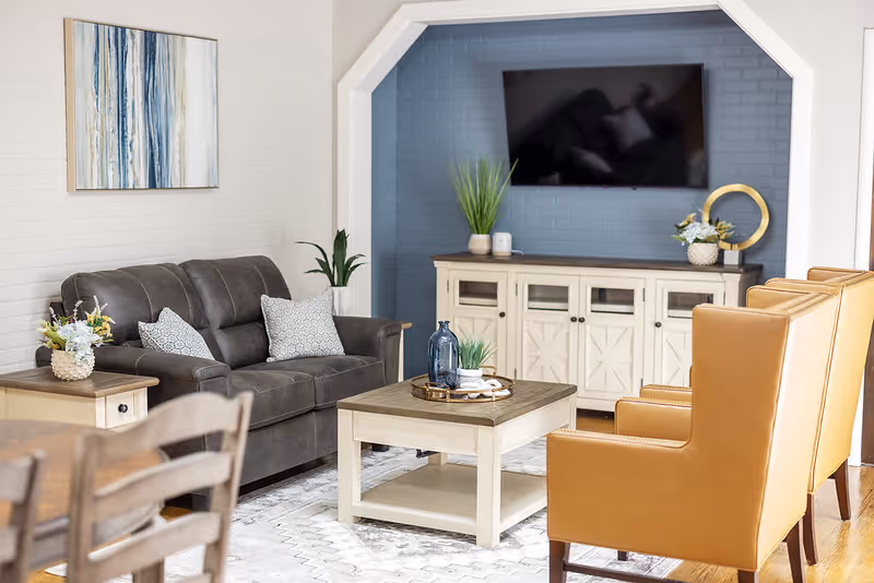 A bright living room with a brown sofa, two tan armchairs, a central coffee table, a media console beneath a wall-mounted TV, and decorative accents.