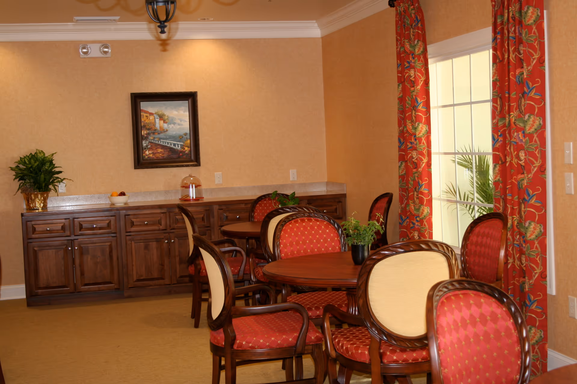 A cozy dining area with round wooden tables and chairs upholstered in red and beige fabric. The room features warm beige walls, a large window with red floral curtains, a wooden cabinet along the back wall with decorative items including a potted plant and a bowl of fruit, and a framed painting hanging above the cabinet.