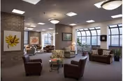 Spacious senior living lounge with chairs, sofas, side tables, artwork, and large windows under ceiling lights.