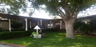 A curved building exterior with a manicured lawn, large tree, and a small white decorative structure.