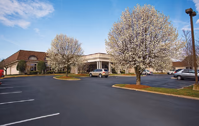 Parking lot with several cars and blooming trees in front of a single-story building under a partly cloudy sky.