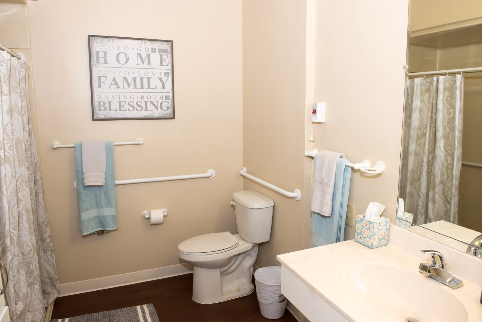 A clean bathroom with a toilet, a sink with a faucet, a mirror, and a shower with a patterned curtain. There are grab bars installed near the toilet and sink, and towels hanging on the bars. A framed sign on the wall reads 'How to go home, how to love family, having both blessing.' A tissue box is placed on the sink counter.