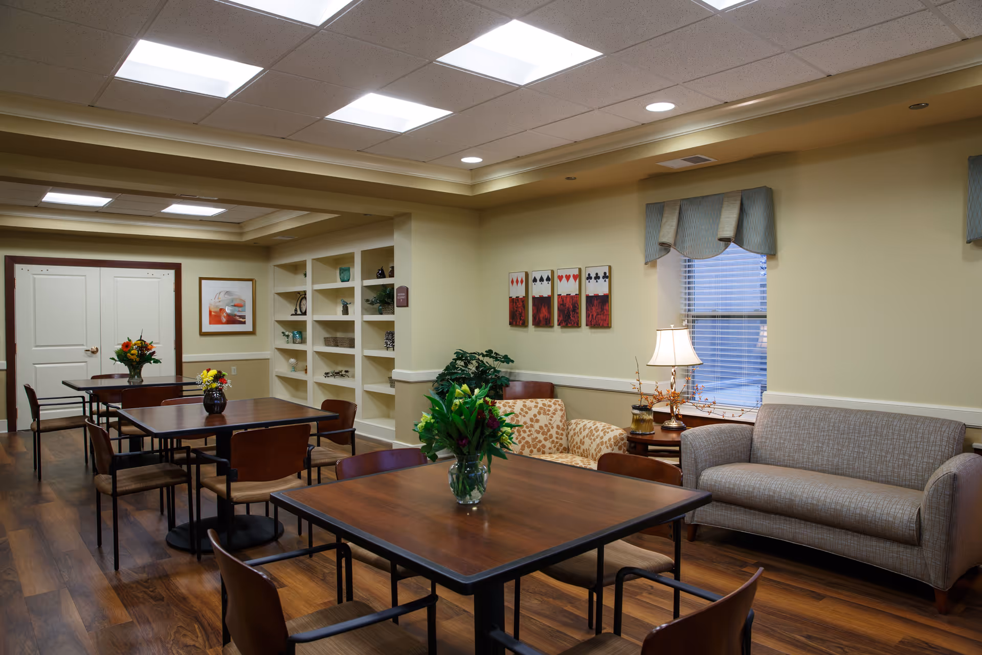 A well-lit senior living common area with wooden tables and chairs arranged for small group seating. There are floral centerpieces on the tables, a beige couch, and a patterned armchair near a window with blinds and a lamp on a side table. The walls are decorated with playing card artwork and shelves holding decorative items. The floor has a wood finish, and the ceiling has recessed lighting panels.