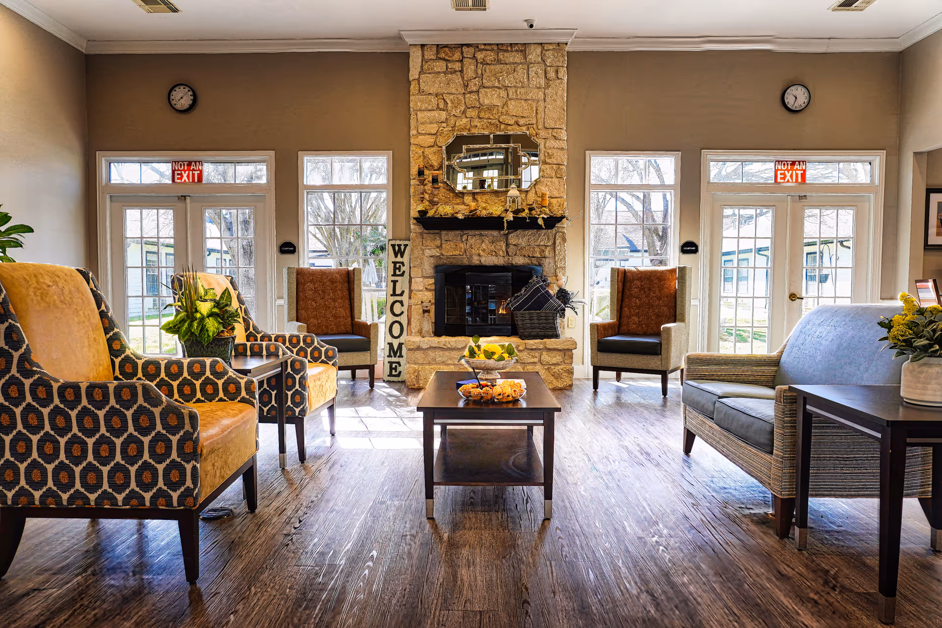 A bright and welcoming living room with wooden flooring, featuring a stone fireplace in the center with a mirror above it and a 'WELCOME' sign leaning against it. The room has multiple armchairs and a sofa arranged around a central coffee table with decorative items. Large windows and glass doors let in natural light, and there are plants and flowers adding a touch of greenery.