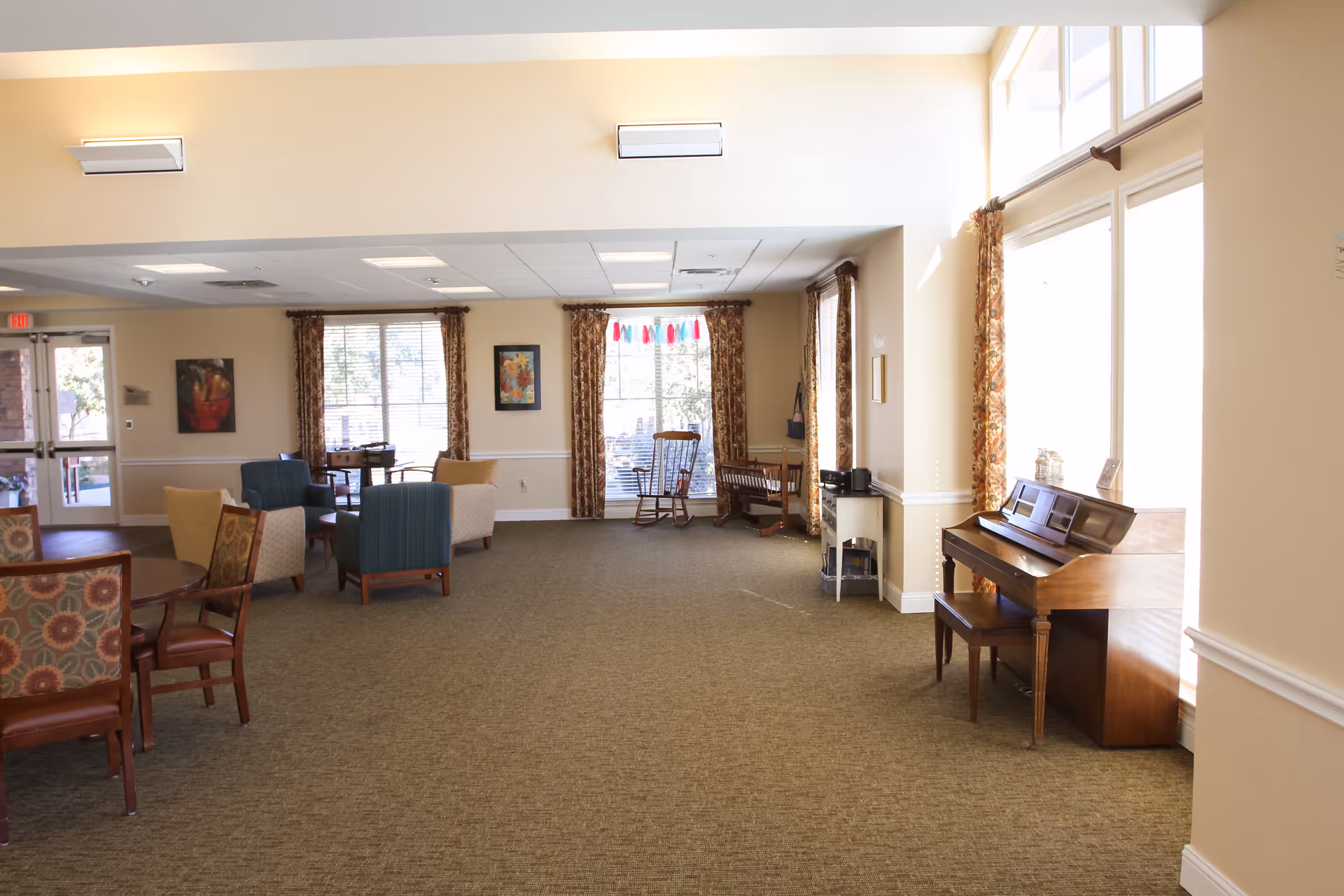 A spacious, well-lit common area in an assisted living facility featuring multiple seating arrangements with chairs and tables, large windows with patterned curtains allowing natural light, a wooden piano with a bench near the windows, and framed artwork on the walls.