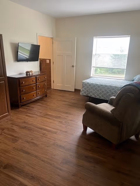 A cozy bedroom with wooden flooring, a large window letting in natural light, a bed with a quilted bedspread, a cushioned armchair, a wooden dresser with drawers, and a wall-mounted flat-screen TV. There is a door leading to another room or bathroom.