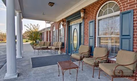 Covered front porch of a brick senior living building with columns, seating, and a teal entry door.