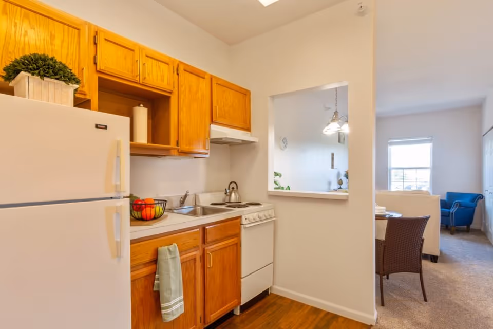A small kitchen area with wooden cabinets, a white refrigerator, a sink, and a stove. A green towel hangs from a cabinet handle, and a bowl of fruit is on the counter. There is a pass-through window opening to a dining and living area with a blue armchair and a window letting in natural light.