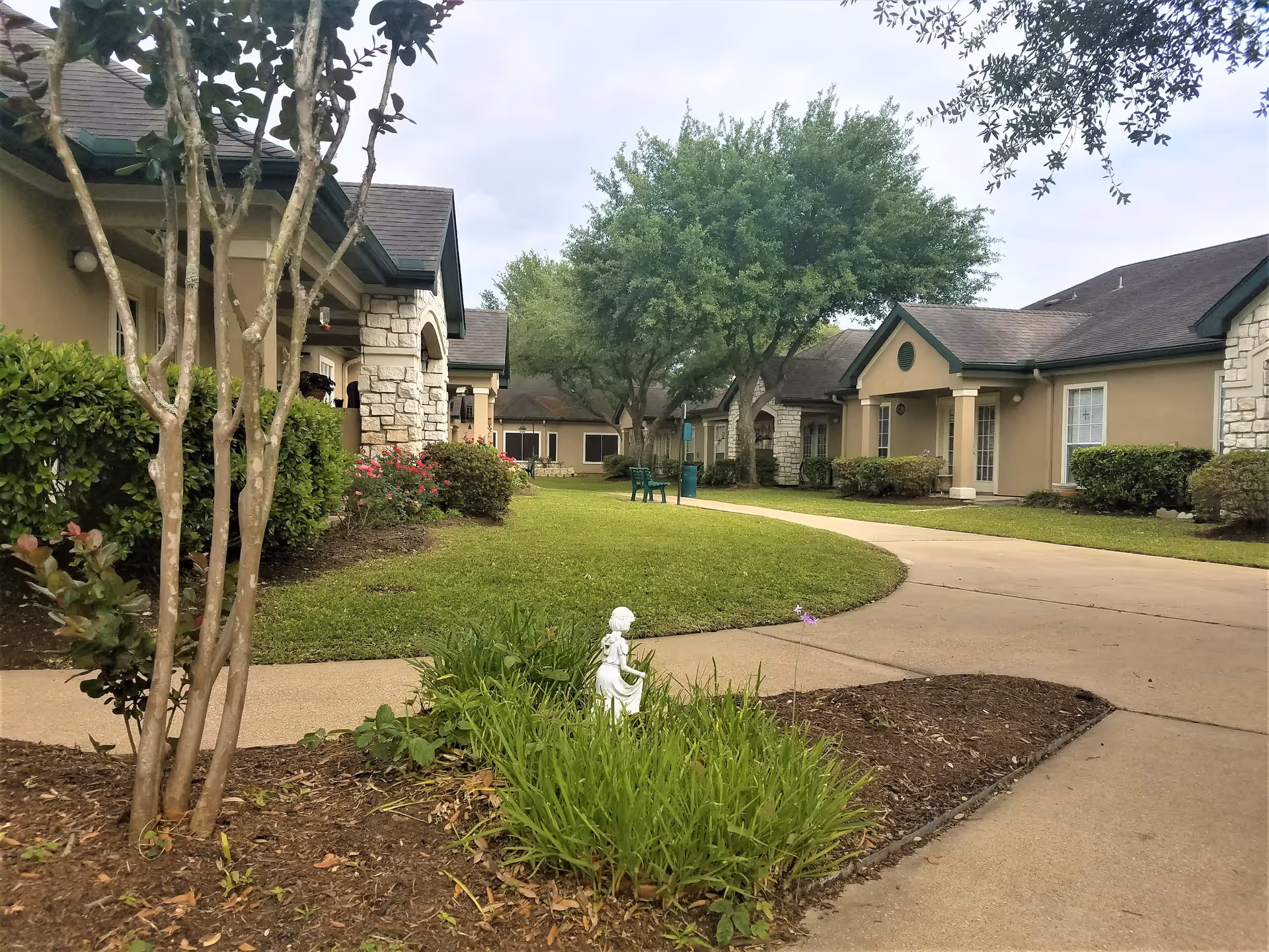 Outdoor view of a senior living facility courtyard with a curved concrete walkway, green grass, bushes, trees, and beige buildings with stone accents and green trim. There is a small white statue in the garden bed near the walkway and two green chairs in the distance.