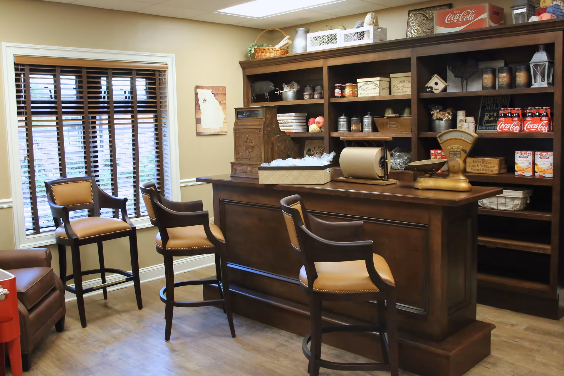 A cozy room with a wooden bar counter and four high chairs with tan cushions. Behind the bar is a large wooden shelving unit filled with various decorative items, canned goods, and vintage containers. A window with wooden blinds is on the left wall, and a small framed map is hung next to it. The floor is wooden, and the overall ambiance is warm and inviting.