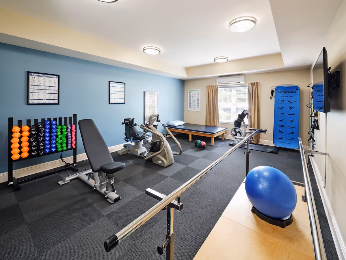 A well-lit exercise room with various fitness equipment including a weight bench, colorful dumbbells on a rack, two stationary exercise bikes, a blue exercise ball, a padded exercise table, and a blue SmartMat with exercise illustrations. The room has a blue accent wall with framed workout posters, a window with beige curtains, and a wall-mounted TV.