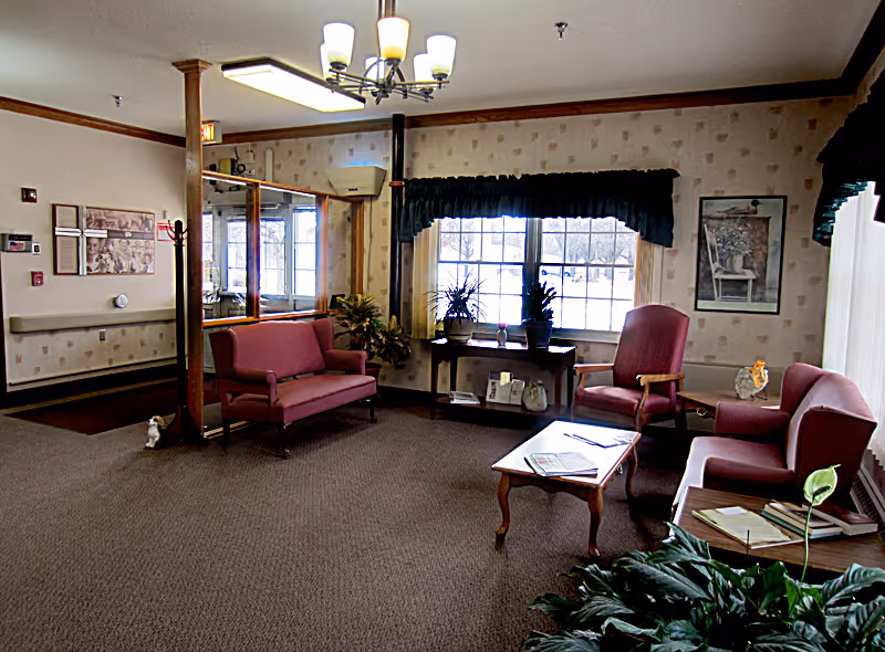 A cozy living room area in a senior living facility with maroon upholstered chairs and a loveseat, a wooden coffee table with magazines, potted plants on a table by the window, and framed artwork on the walls. The room has carpeted flooring, patterned wallpaper, and a ceiling light fixture.
