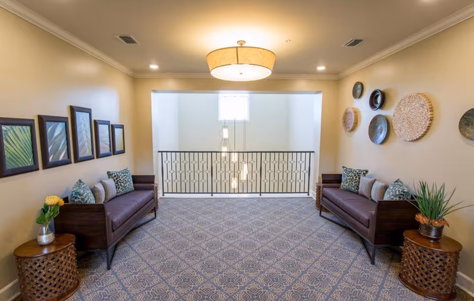 A cozy sitting area with two dark wooden sofas facing each other, each adorned with patterned cushions. Between the sofas are round wooden side tables with decorative plants and flowers. The walls are decorated with framed artwork on one side and circular woven wall hangings on the other. The area is carpeted with a patterned rug and illuminated by a ceiling light fixture. A railing overlooks a lower level with hanging pendant lights and a window providing natural light.