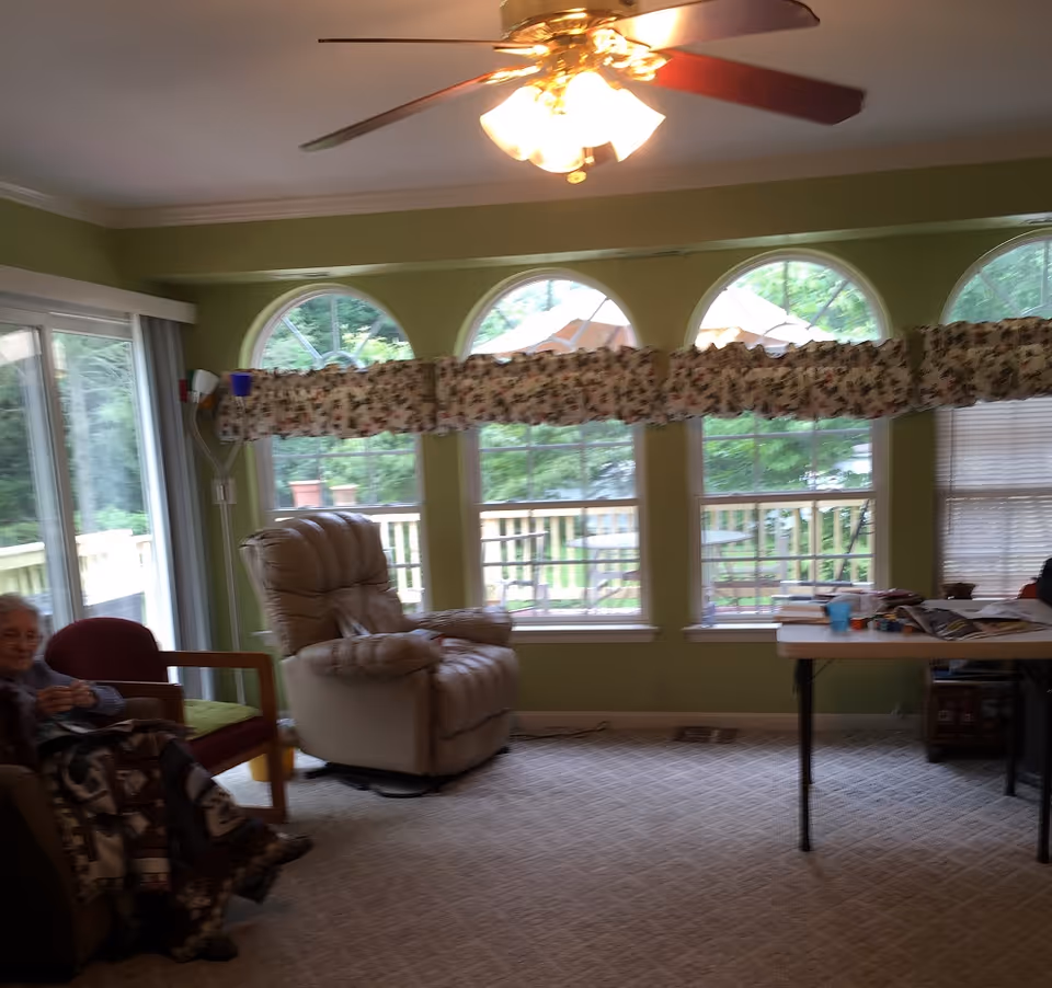 A living room area with green walls and large arched windows covered partially by floral valances. There is a beige recliner chair, a red armchair with a person sitting on it covered with a blanket, a table with various items on it, and a ceiling fan with lights. Outside the windows, a wooden deck and green trees are visible.