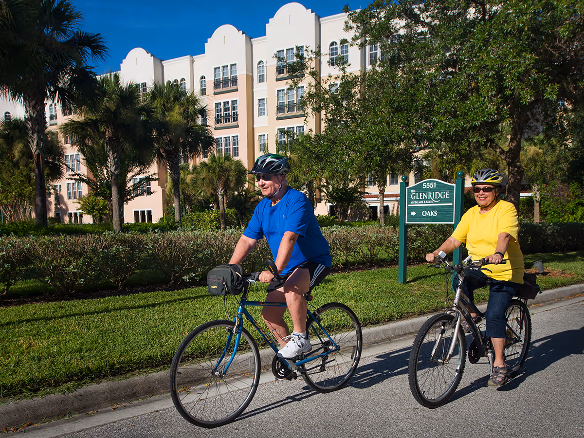 Two older adults wearing helmets ride bicycles past a sign for The Glenridge in front of a palm-lined, multi-story senior living building.