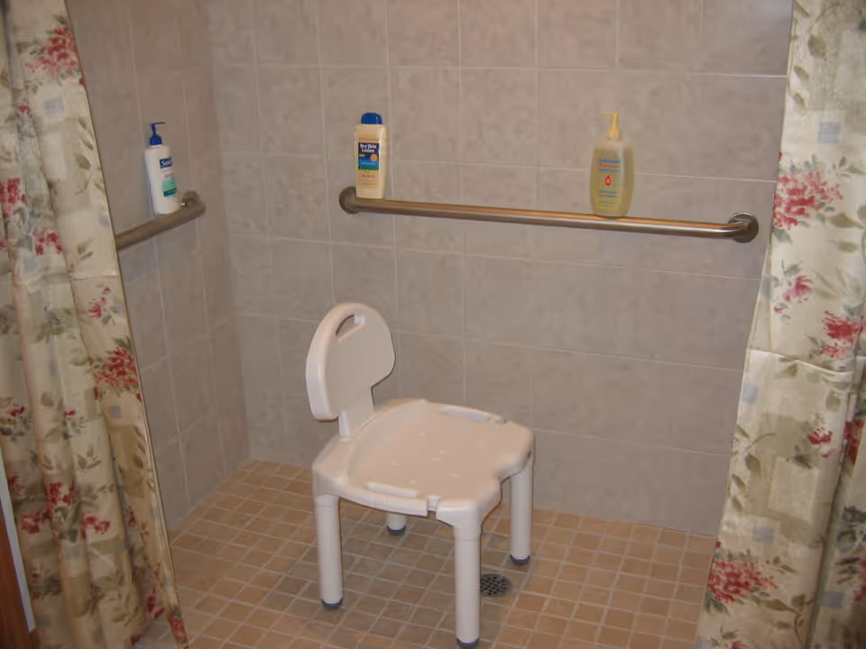 A tiled shower area with a white plastic shower chair with a backrest. There are two metal grab bars mounted on the tiled walls, one horizontal and one vertical. Three bottles of lotion or soap are placed on the grab bars. Floral patterned shower curtains are partially visible on both sides.