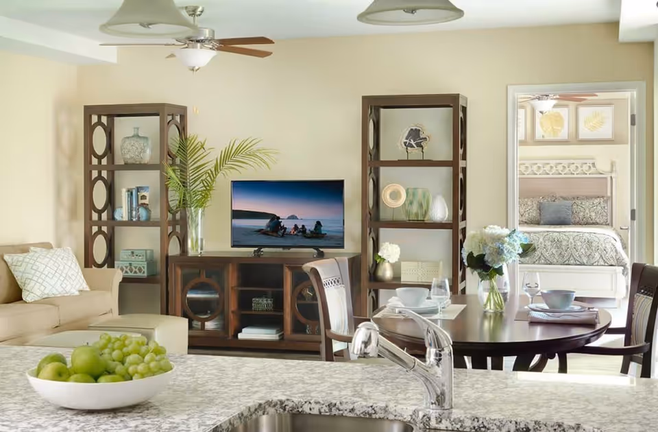 View of a cozy senior living community interior showing a living room with a beige sofa, two wooden shelving units flanking a TV on a wooden stand, a dining table set with dishes and flowers, and a bedroom visible through an open doorway. The foreground features a kitchen counter with a bowl of green apples and grapes and a modern faucet.