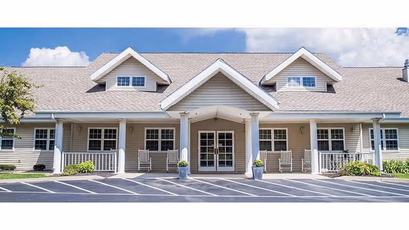 Front entrance of a single-story senior living building with a covered porch, columns, rocking chairs, and parking spaces.
