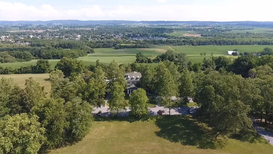 Aerial view of a large country estate house surrounded by trees, a front lawn, and rolling farmland in the distance.
