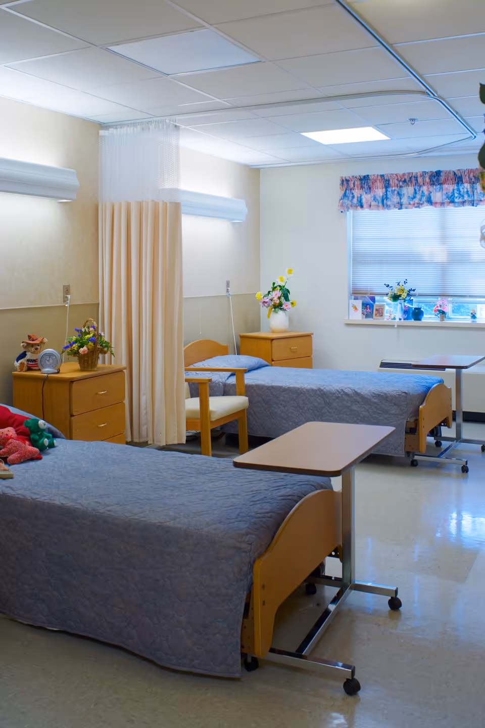 A shared bedroom in a senior living facility with two single beds covered in gray quilts, each accompanied by a wooden nightstand with flowers and personal items. There is a beige privacy curtain between the beds, a wooden chair, and a window with floral valance letting in natural light.