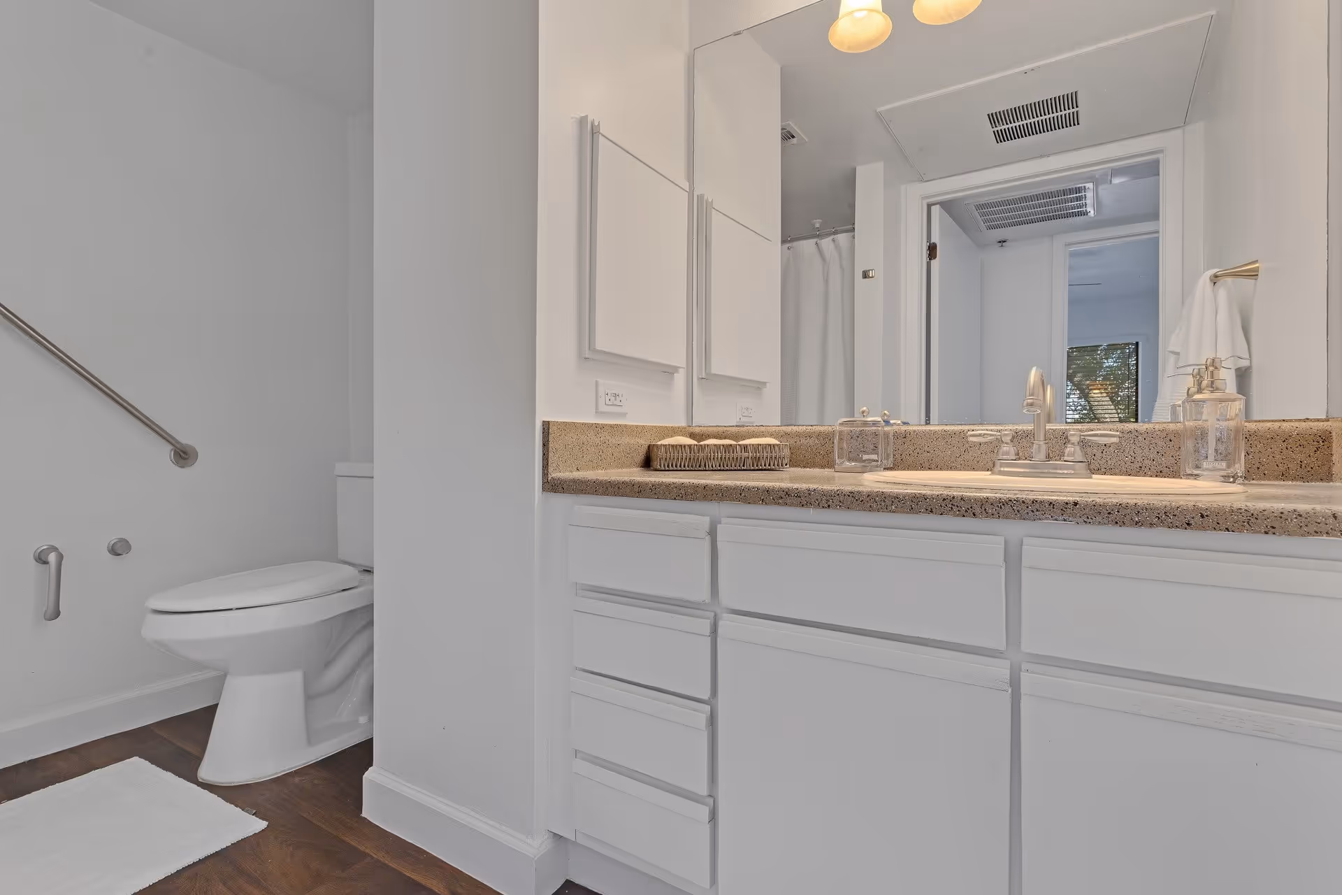 A clean and modern bathroom featuring a white toilet with a grab bar on the wall, a white vanity with multiple drawers, a speckled countertop with a sink, soap dispenser, and a basket with towels. A large mirror is mounted above the vanity, reflecting a doorway and part of another room.