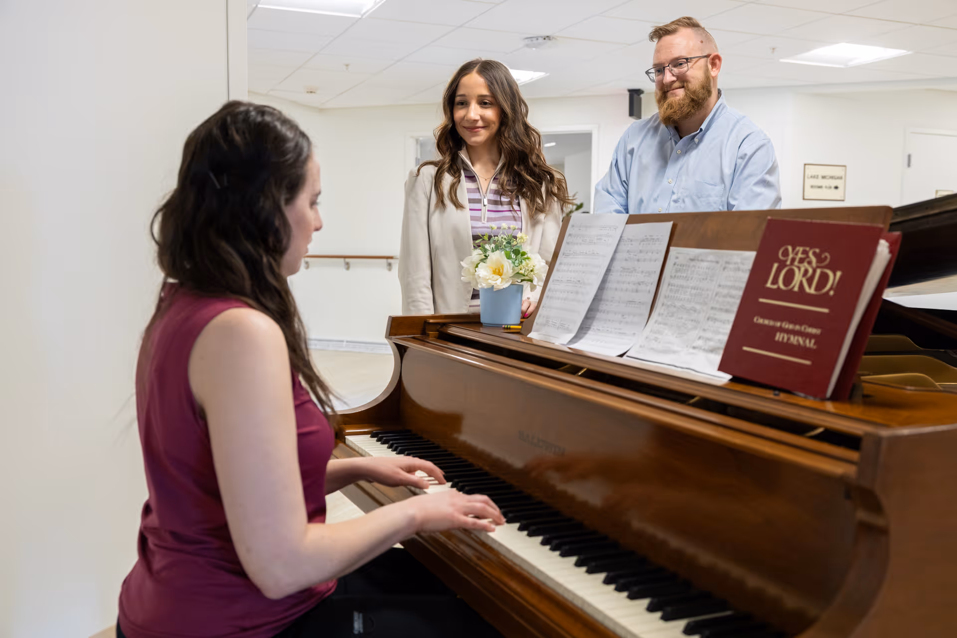 A woman in a sleeveless maroon top is playing a grand piano with sheet music and a hymnal titled 'YES LORD!' on the music stand. Two people, a woman in a white blazer and a man with glasses and a beard wearing a light blue shirt, stand nearby watching and smiling. The setting appears to be an indoor common area with white walls and ceiling lights.