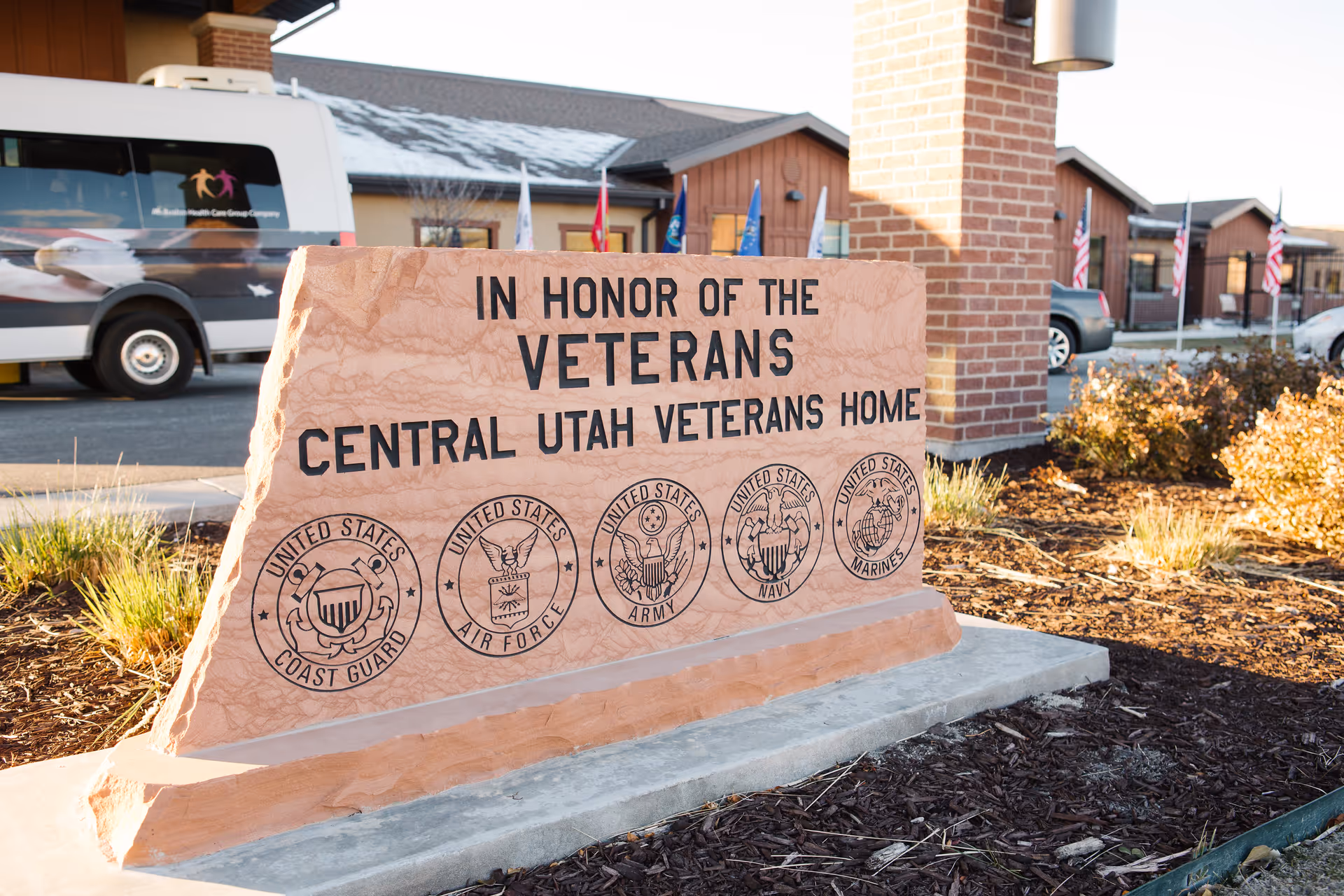 A large stone monument outside the Central Utah Veterans Home with inscriptions honoring veterans. The monument features the text 'IN HONOR OF THE VETERANS CENTRAL UTAH VETERANS HOME' and displays the seals of the United States Coast Guard, Air Force, Army, Navy, and Marines. In the background, there is a white van, a brick pillar, several flags, and parts of the facility buildings.