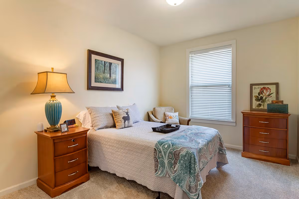 A cozy bedroom with a neatly made bed featuring a white quilt and a patterned throw blanket. There are two pillows, one with a deer design and another with the word 'Home'. A wooden nightstand with a blue lamp, picture frames, and a small device is beside the bed. Across the room, there is a wooden dresser with decorative items and a framed floral picture. A window with white blinds lets in natural light, and a comfortable armchair with a decorative pillow is placed near the window. The walls are painted light beige, and the carpet is light-colored.