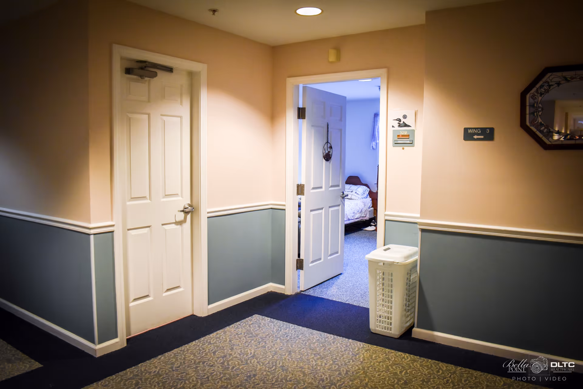 Interior hallway of a senior living facility with beige and blue walls, a closed white door on the left, and an open door leading to a bedroom with a bed visible. A white laundry basket is placed near the open door. A sign on the wall indicates Wing 3, and a decorative mirror hangs on the right wall.