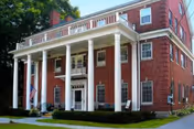 Exterior front view of a large red brick building with white columns supporting a balcony above the entrance. The building has multiple windows and an American flag displayed near the entrance. There is a well-maintained lawn and some greenery around the building.