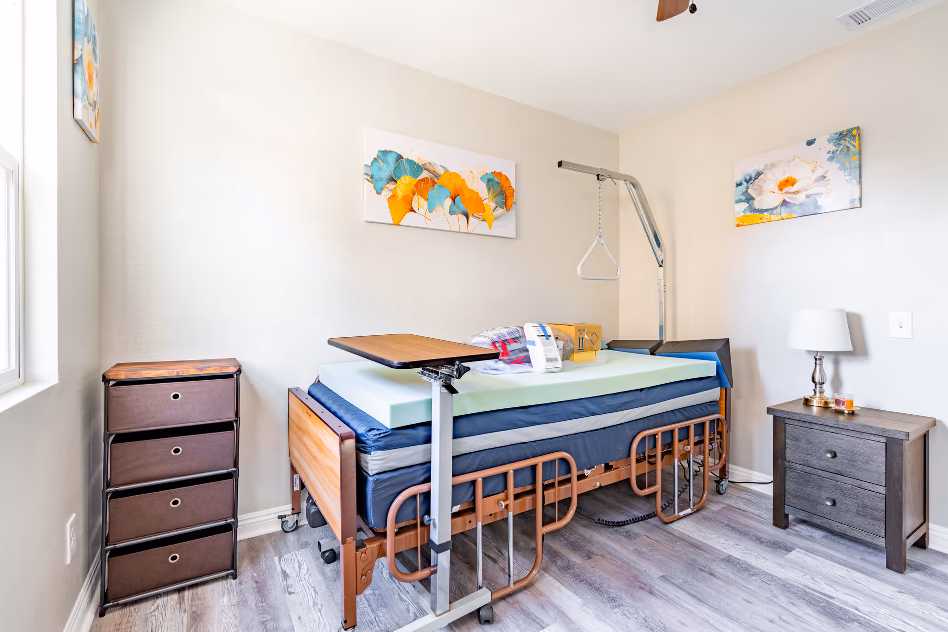 A small bedroom with a hospital-style bed featuring side rails and an overhead trapeze bar. The bed has a green mattress topper and a wooden overbed table. There is a dark brown chest of drawers on the left and a matching nightstand with a lamp and small items on the right. The walls are light-colored and decorated with two colorful floral paintings. The floor is a light wood laminate.