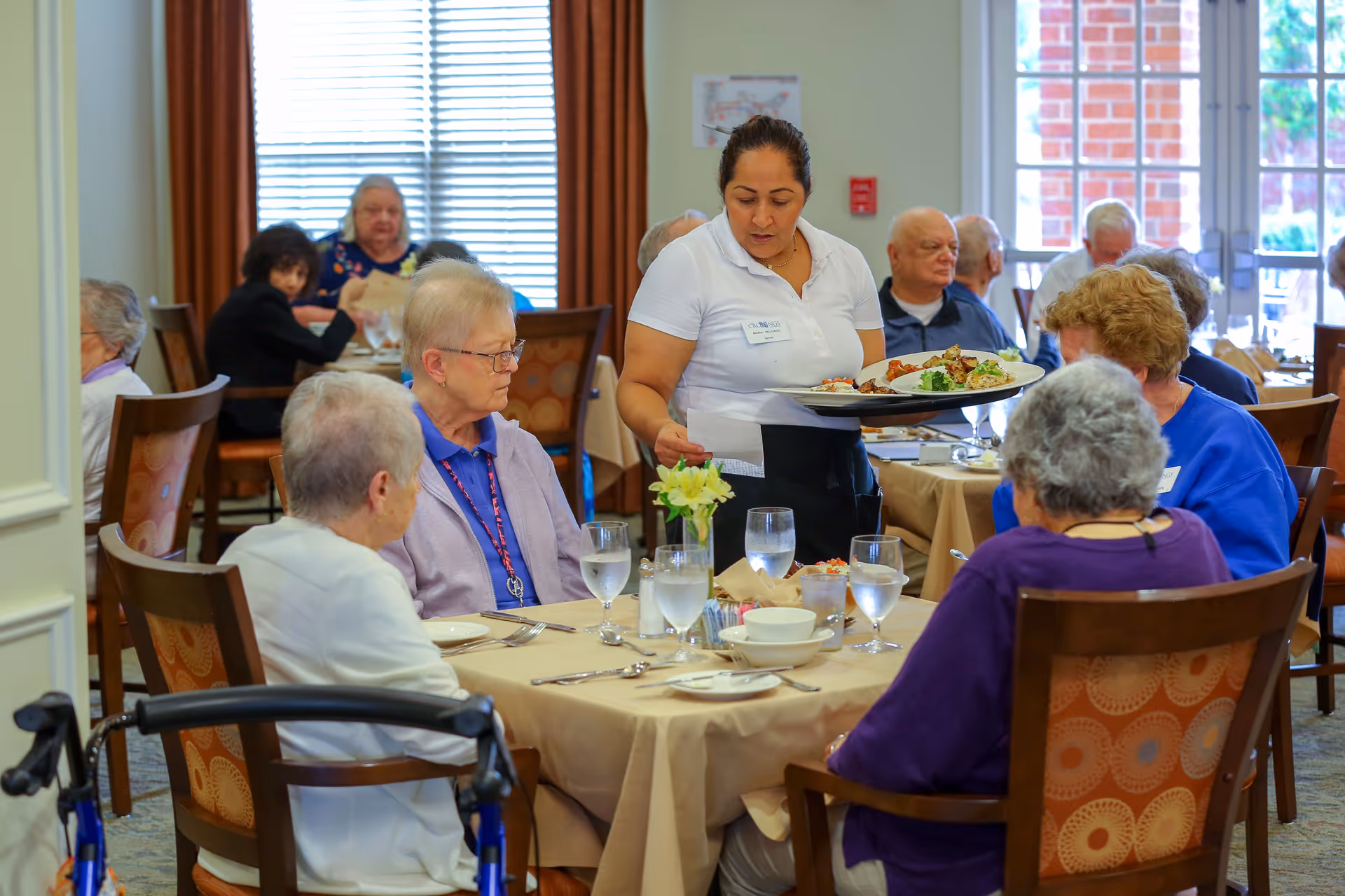 A dining room scene at a senior living facility where elderly residents are seated around tables with beige tablecloths. A waitress is serving a plate of food to one of the tables. The room has large windows with natural light and brown curtains, and the tables are set with glasses of water, bowls, and silverware.