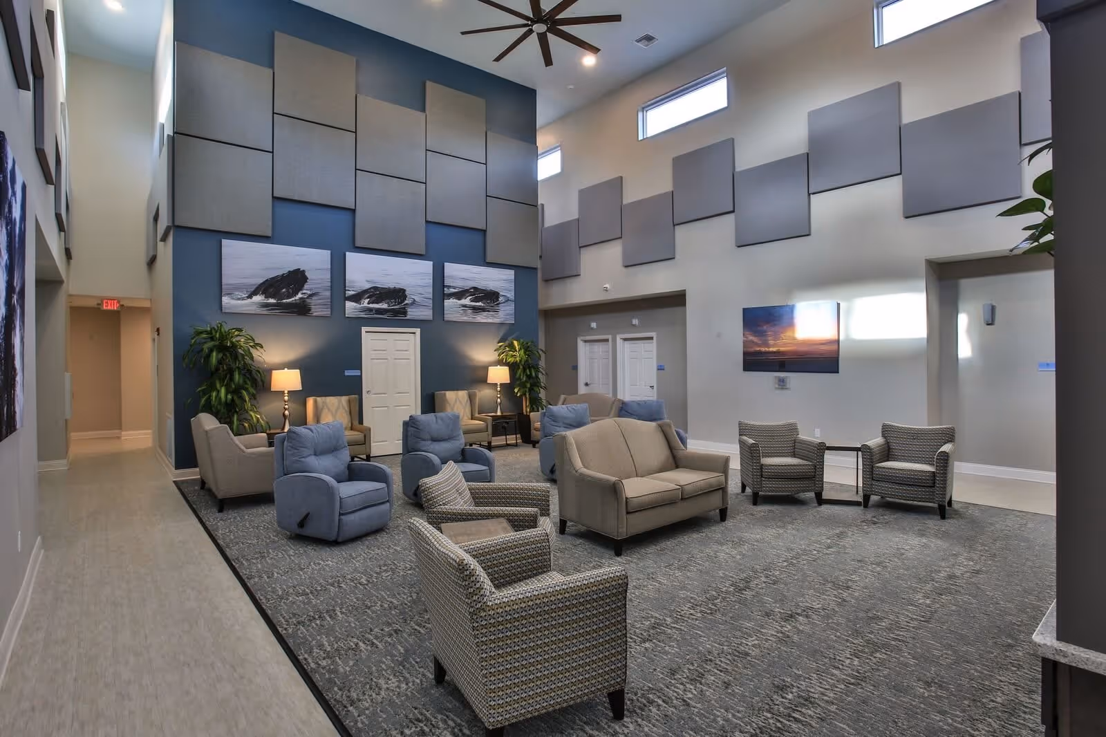 A spacious and well-lit common area with multiple armchairs and sofas arranged on a large carpet. The walls are decorated with modern art and acoustic panels. There are tall windows near the ceiling allowing natural light to enter, and ceiling fans are installed above. Two potted plants and table lamps add to the cozy atmosphere.