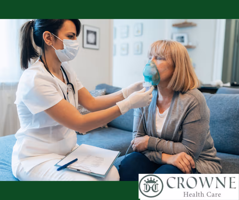 A healthcare professional wearing a mask and gloves assists an elderly woman sitting on a couch by helping her use an oxygen mask. The healthcare worker has a clipboard on her lap.