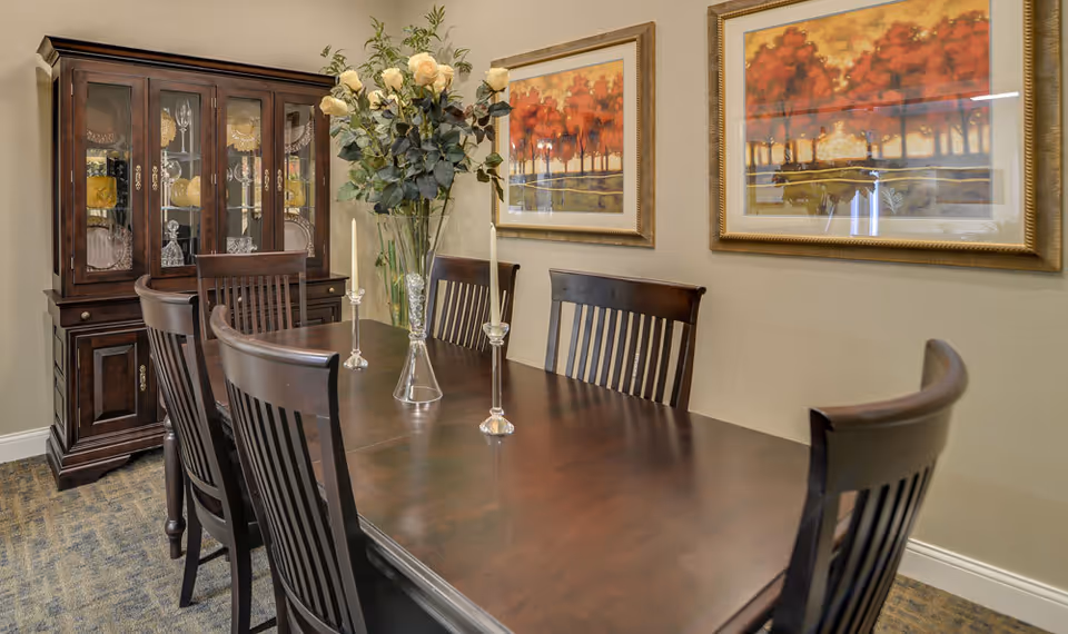 A dining room with a dark wooden table and six matching chairs. On the table are three tall candle holders and a vase with white flowers. Behind the table is a wooden china cabinet displaying glassware and plates. Two framed paintings of autumn trees hang on the beige wall.