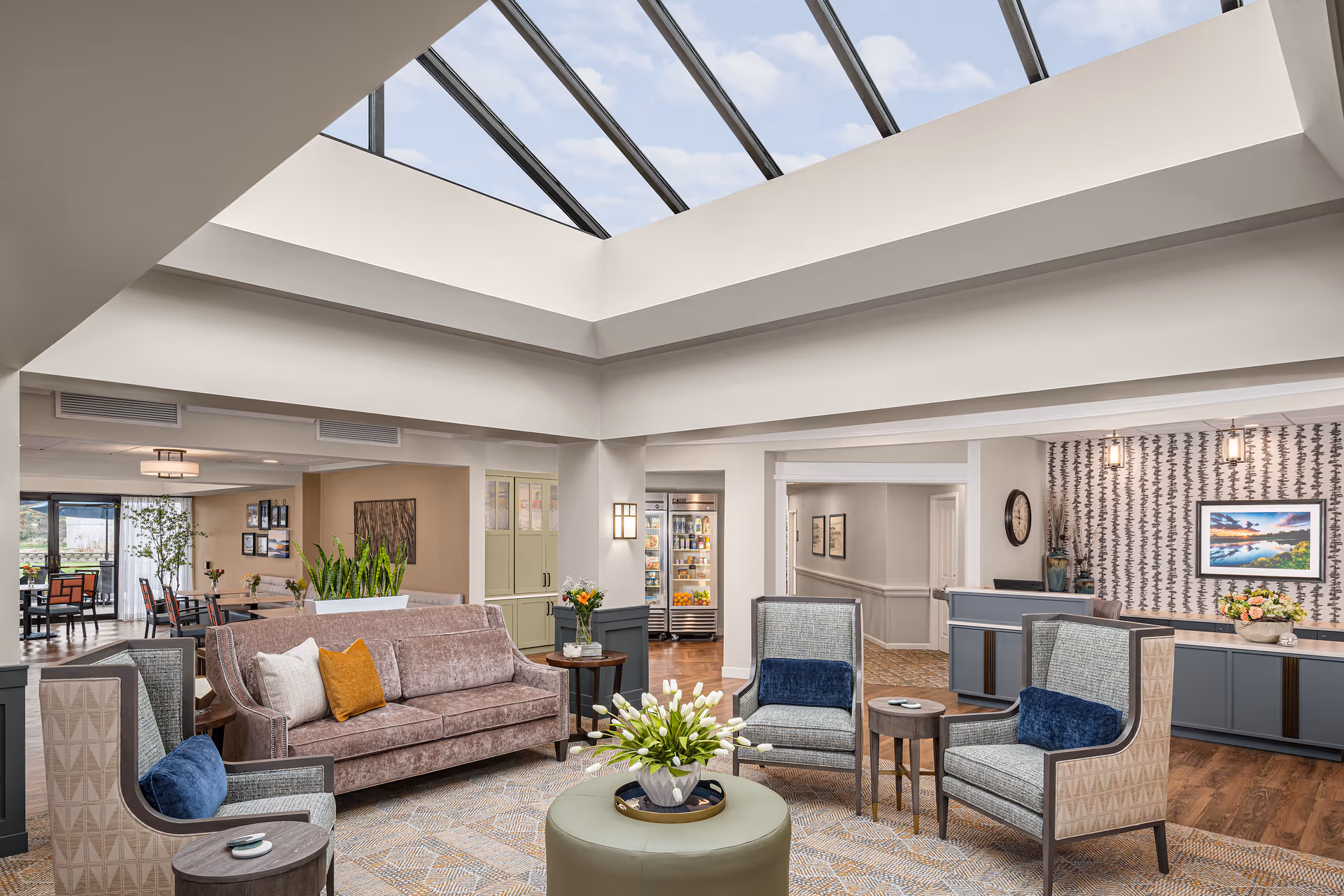 Bright communal lounge at Harbour House with a large skylight, upholstered sofa and chairs arranged around a coffee table, and a reception desk and dining area visible in the background.