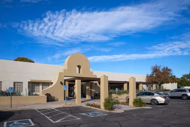 Front entrance of a single-story beige rehabilitation facility with an arched porte-cochere, handicapped parking spaces and cars under a blue sky.