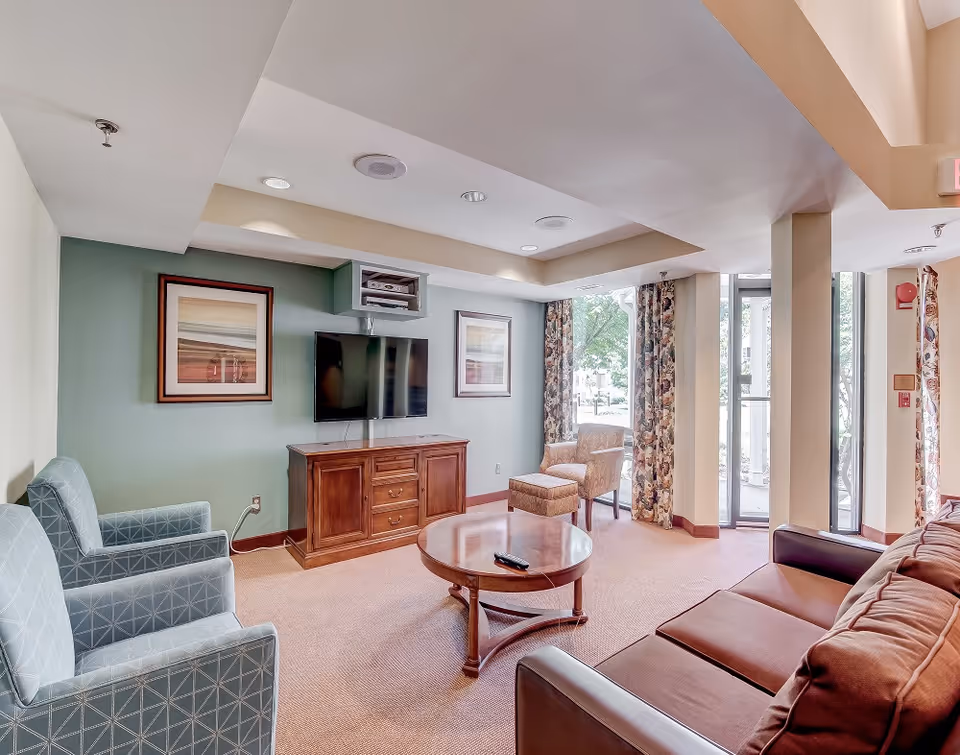 A bright common lounge with sofas and armchairs arranged around a coffee table, a wall-mounted TV on a wooden cabinet, and large windows with patterned curtains.