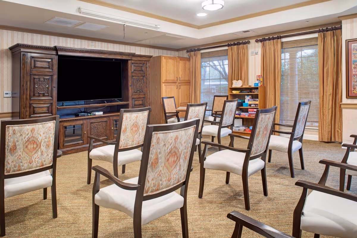 A senior living common room with upholstered chairs arranged facing a large wooden entertainment center and TV.