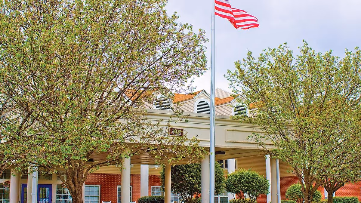 Entrance of a senior living building with white columns, an American flag on a pole, and leafy trees in front.