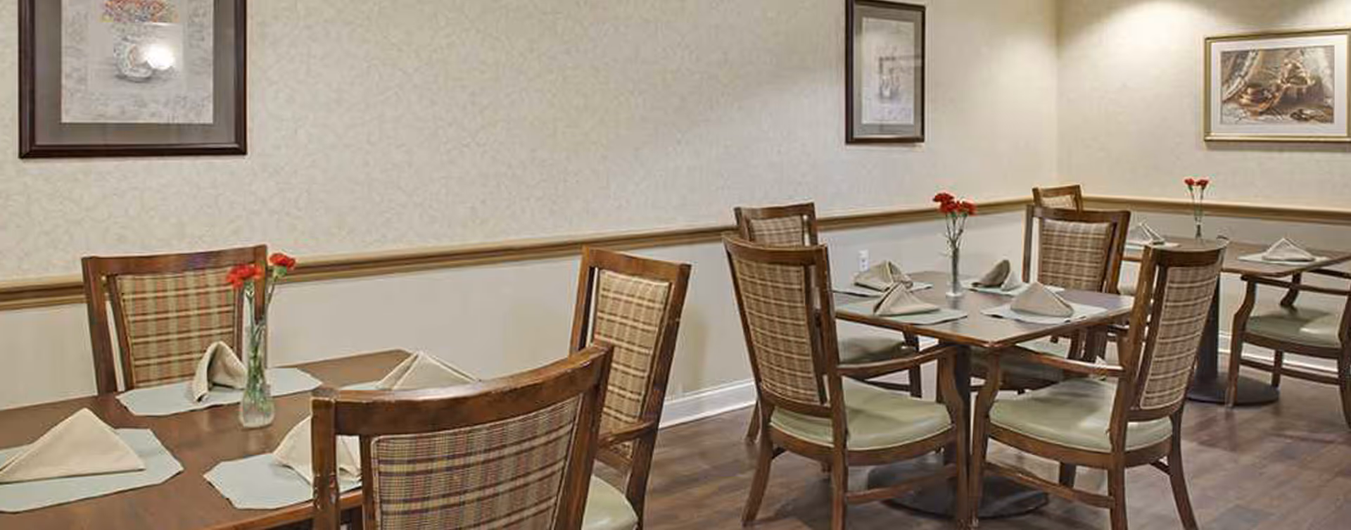 Dining room with wooden tables and chairs set with folded napkins and small vases of red flowers.