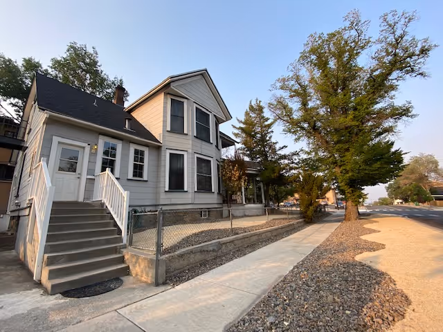 Exterior view of a two-story residential building with white siding and black roof shingles. There is a concrete staircase with white railings leading to a door on the left side. The building is bordered by a chain-link fence and a sidewalk runs alongside it. Trees and a street are visible on the right side under a clear sky.