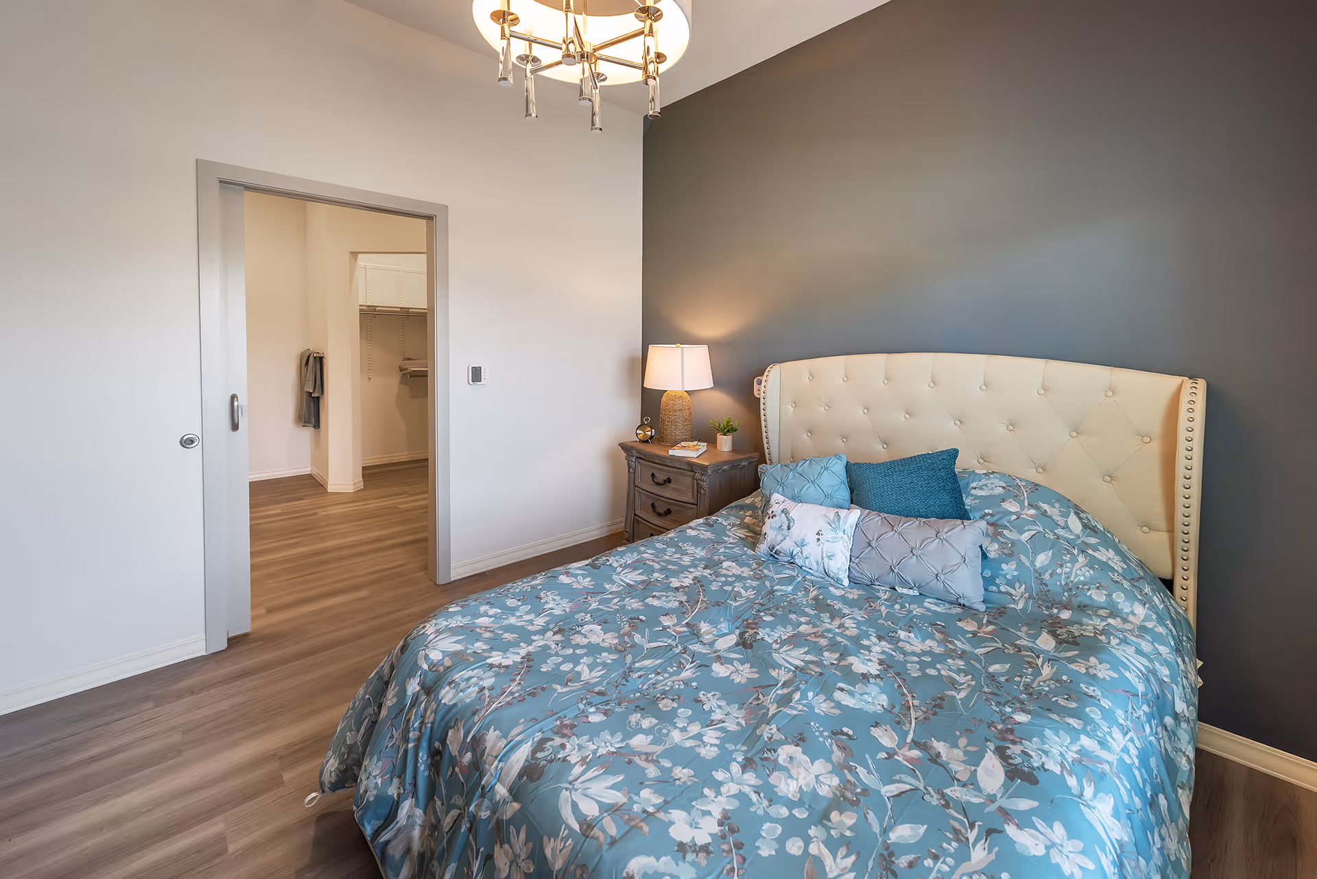 Neatly decorated bedroom with an upholstered headboard, floral bedspread, nightstand and lamp, and an open doorway to a walk-in closet.
