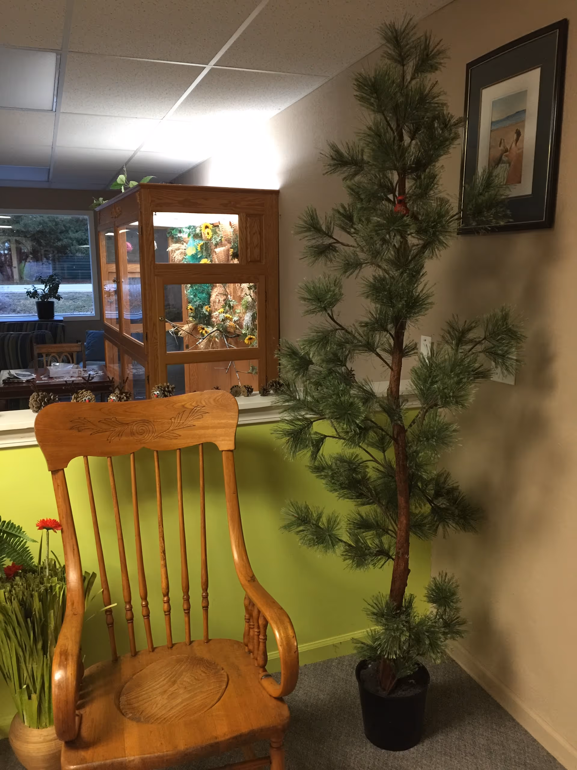 Wooden rocking chair beside a tall potted artificial pine tree in an interior sitting area with a display case and framed art in the background.