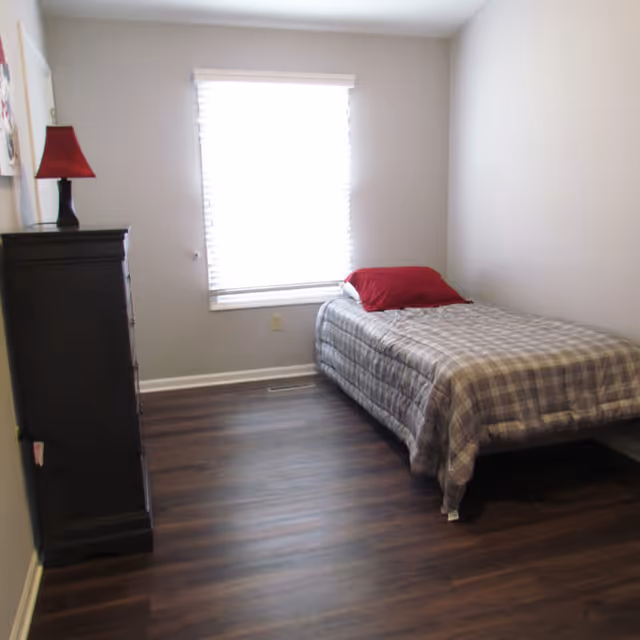 Small bedroom with a single bed against the right wall, a dresser with a red lamp, and a window with closed blinds.
