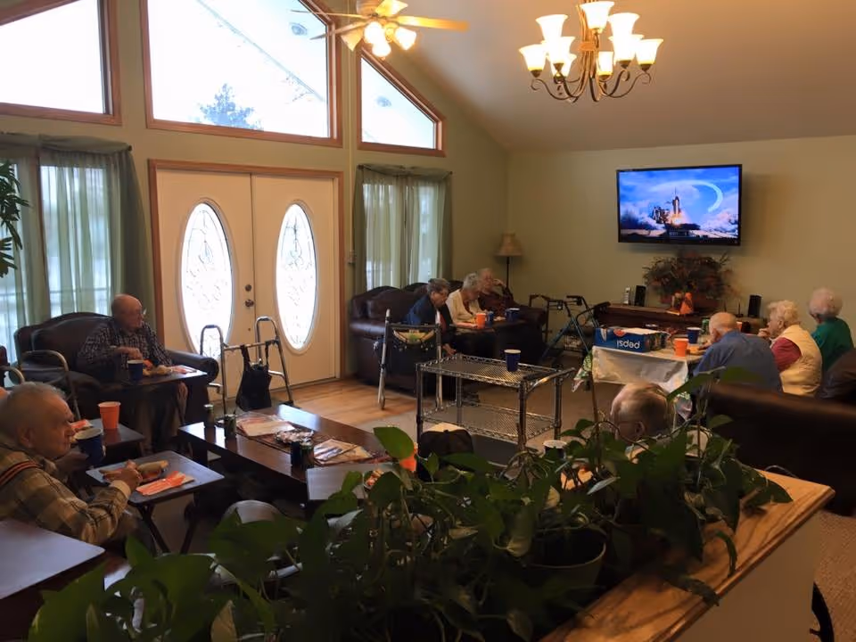 A common living room in an assisted living facility with seniors seated around tables watching a wall-mounted TV.